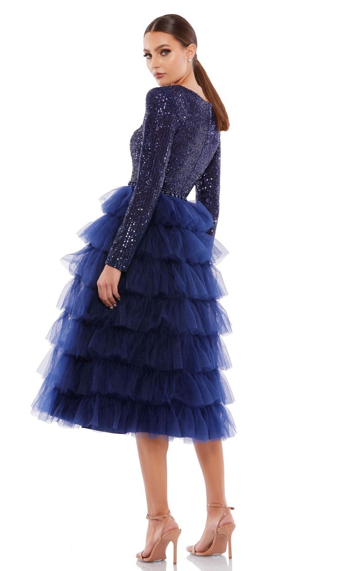 Woman wearing the Mac Duggal 26299 knee-length wedding guest dress in navy blue with long sleeves, sequins, and a layered tulle skirt, posing with her back turned and head over her shoulder.